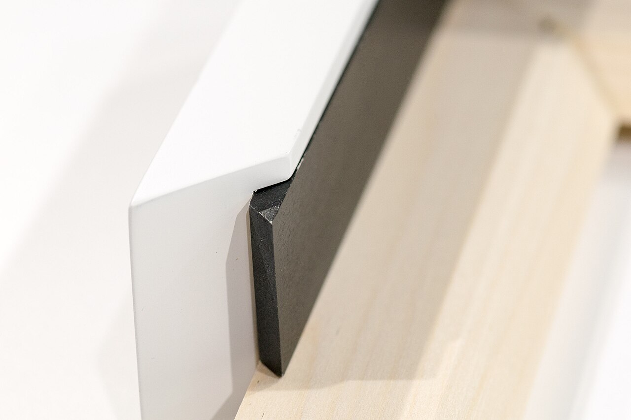 Close-up of the corner of a picture frame in cross section: a white outer moulding, a black glass spacer and the pale wooden subframe behind